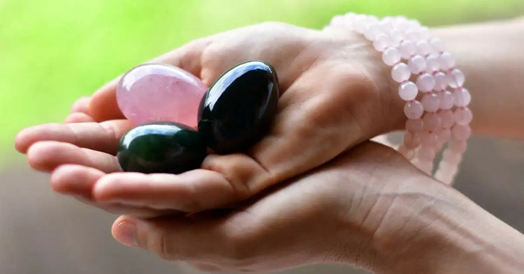Une main tient trois œufs en pierre gemme lisses et polis — quartz rose rose, obsidienne noire et jade vert foncé — dans un cadre naturel.